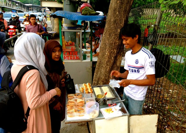 Two girls buy apem, a tasty snack, in the middle of a busy sidewalk in central Jakarta.